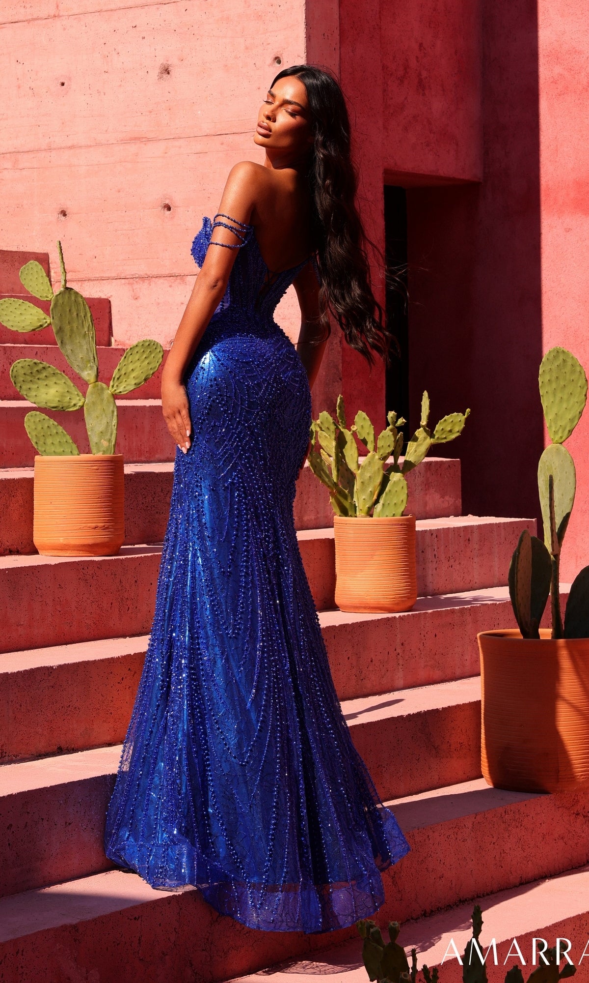 Wearing the Amarra 89610, a sparkling royal blue long prom dress with an off-the-shoulder design and corset bodice, a woman stands on terracotta steps adorned with potted cacti beside a pink textured wall, looking over her shoulder.